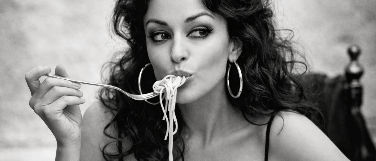 Eine Frau in einem schwarzen Kleid isst Spaghetti mit einer Gabel. Das Foto ist schwarz-weiß.