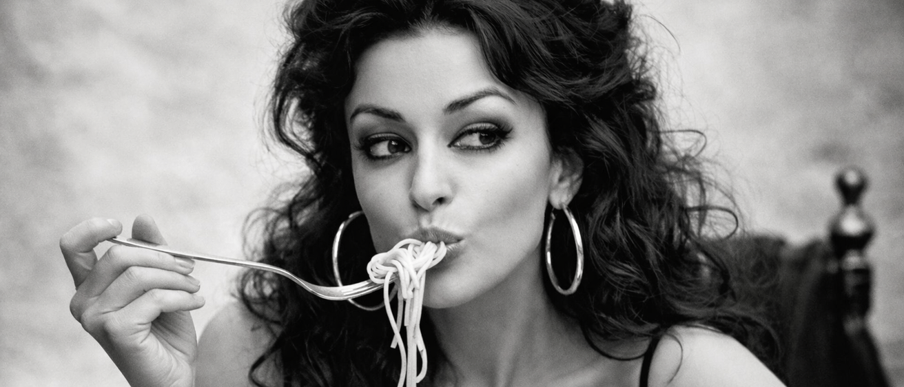 Eine Frau isst Spaghetti mit einer Gabel, während sie sitzt. Sie hat lockiges Haar und trägt Hoop-Ohrringe. Das Bild ist in Schwarz-Weiß.