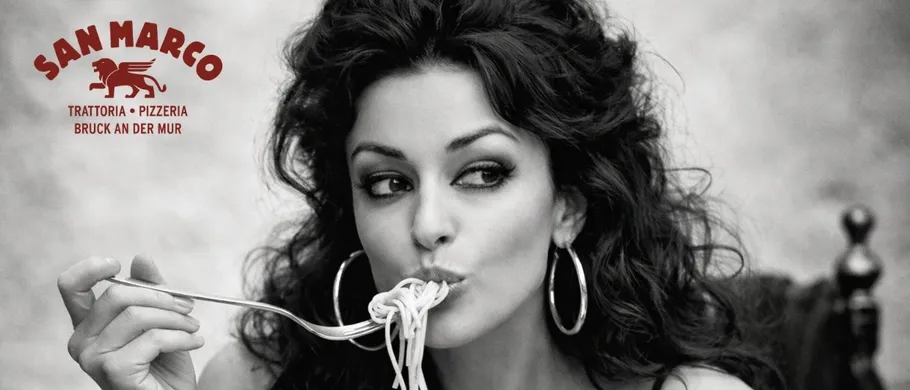 Ein Schwarz-Weiß-Foto zeigt eine Frau mit lockigen Haaren, die Spaghetti mit einer Gabel isst. Das Bild enthält ein Restaurantlogo und Text.