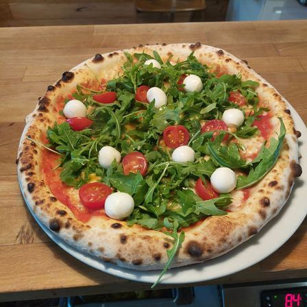Eine Pizza mit Rucola, Mozzarella und Kirschtomaten liegt auf einem weißen Teller auf einem Holztisch.