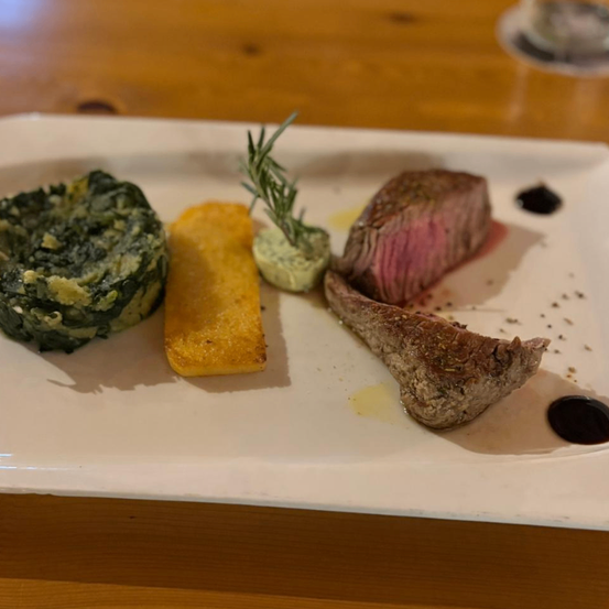 Ein weißer Teller enthält ein Steak, ein grünes Gemüse und ein Stück gebratenes Brot, mit Rosmarin und einer grünen Sauce.