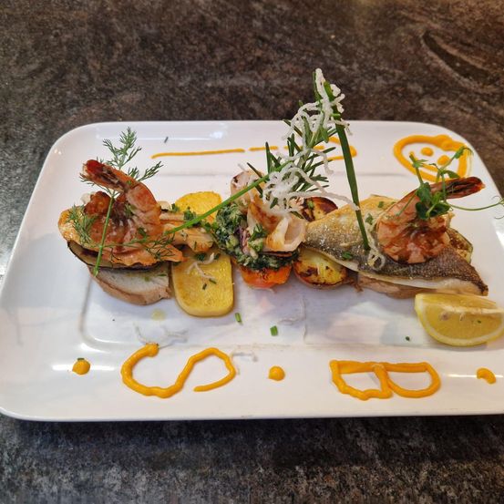 Ein Teller mit gegrilltem Fisch, Garnelen und Gemüse, garniert mit Kräutern und Zitronenstücken, präsentiert auf einem weißen Teller mit dekorativen orangefarbenen Schwüngen.