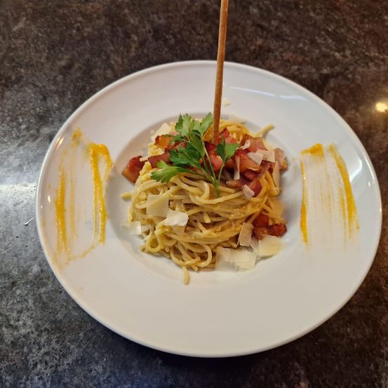 Ein Teller mit Spaghetti, Speck, Käse und Petersilie auf einem Marmortisch, mit einem Zahnstocher, der herausragt.