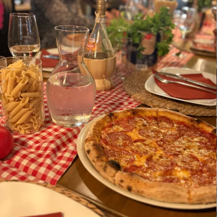 Eine Pizza mit Peperoni und Käse steht auf einem Tisch mit einer rot-weiß karierten Tischdecke. Im Hintergrund befinden sich Pasta, Wein und eine Pflanze.