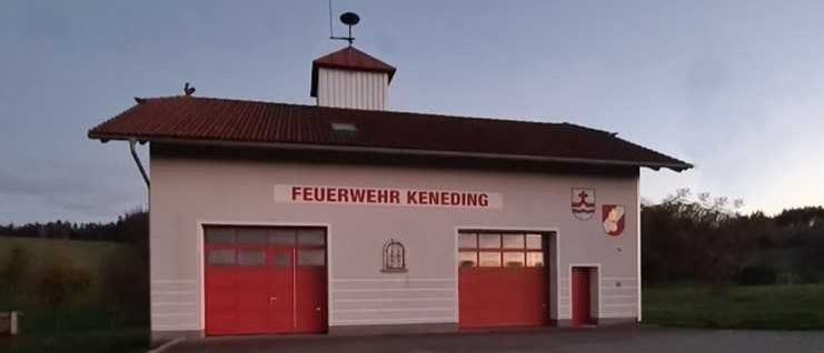 Ein weißes Gebäude mit roten Türen und einem roten Dach trägt die Aufschrift 'FEUERWEHR KENEDING' mit einem Logo an der Seite.