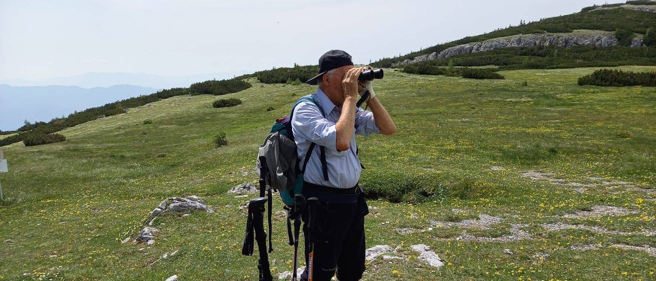 Ein Wanderer steht auf einem grasbewachsenen Hügel mit Fernglas und schaut in die Ferne. Er trägt einen Hut, einen Rucksack und Wanderschuhe.