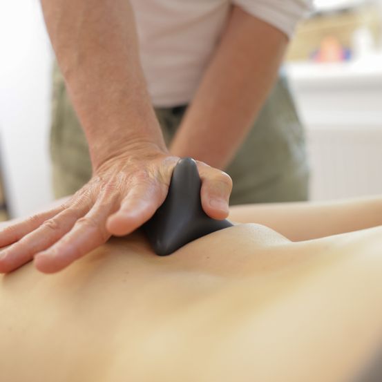 Eine Person massiert eine andere Person mit einem schwarzen Werkzeug auf dem Rücken in einem Raum.