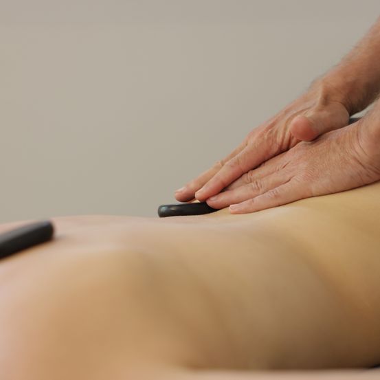 Eine Person liegt auf einem Massagetisch und erhält eine Massage. Die Masseurin verwendet ein schwarzes Massagewerkzeug auf dem Rücken der Person.