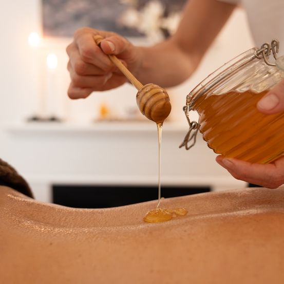 Eine Person gießt Honig mit einem Holzlöffel auf den Rücken einer anderen Person. Der Honig tropft vom Löffel.