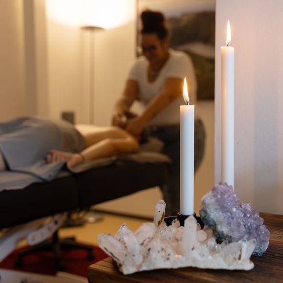 Eine Person liegt auf einem Massagetisch, während ein Therapeut an ihr arbeitet. Zwei brennende Kerzen und Kristalle stehen auf einem Tisch. Der Raum hat ein Bild an der Wand und eine Lampe.