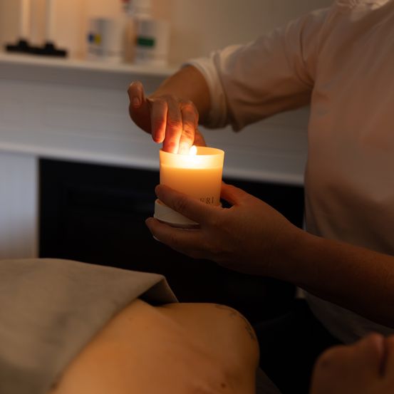 Eine Person in einem weißen Hemd zündet eine Kerze in einem schwach beleuchteten Raum an. Die Kerze steht auf einem silbernen Halter.