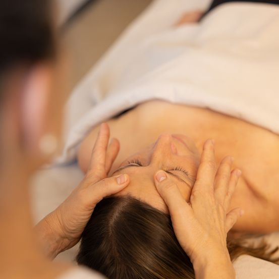 Eine Frau liegt auf einem Bett, mit geschlossenen Augen, und erhält eine Gesichtsmassage von einer Person mit den Händen auf ihrem Gesicht.