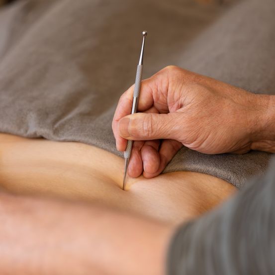Eine Hand hält ein metallisches Werkzeug gegen einen nackten Bauch, was auf eine medizinische oder Akupunktur-Prozedur hindeutet.