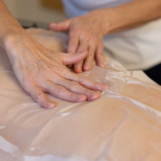 Eine Person massiert den Rücken einer anderen Person, die mit einer Plastikfolie bedeckt ist. Die Hände der Person liegen auf dem Rücken und üben Druck aus.