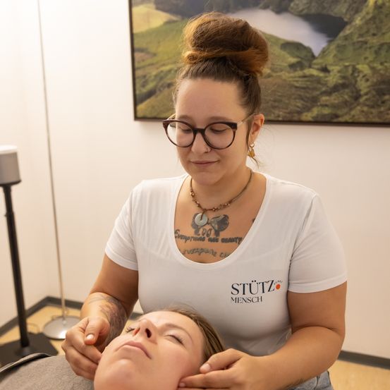 Eine Frau mit Tätowierungen massiert die Stirn einer anderen Frau mit Brille in einem weißen Raum.