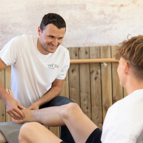 Ein Mann in einem weißen STUTZ MENSCH T-Shirt führt eine Fußmassage bei einem Jungen in einem weißen T-Shirt durch, mit einer Holzwand im Hintergrund.