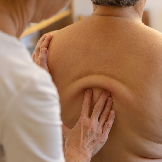 Eine Person in einem weißen T-Shirt massiert die Schulter einer anderen Person, hält sie mit einer Hand und drückt sie mit der anderen.