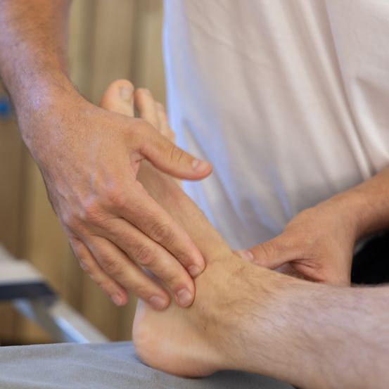 Eine Person erhält eine Fußmassage von einer anderen Person, die ein weißes Hemd trägt. Die Füße der Person liegen auf einer grauen Oberfläche, und der Masseur hält die Füße mit beiden Händen.