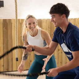 Ein Mann und eine Frau trainieren mit einem Battle Rope in einem Fitnessstudio, lächeln und schauen sich an. Die Frau hat Tattoos an den Armen.