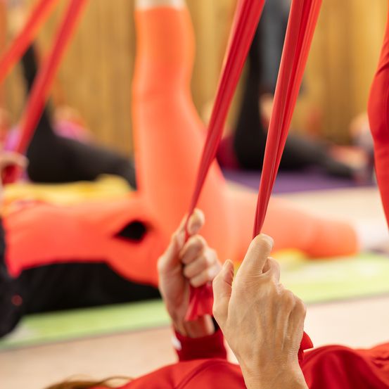 Eine Gruppe von Menschen praktiziert Yoga mit roten Yoga-Riemen. Sie liegen auf Yogamatten auf dem Boden, mit erhobenen Beinen. Einige sind im Hintergrund verschwommen.