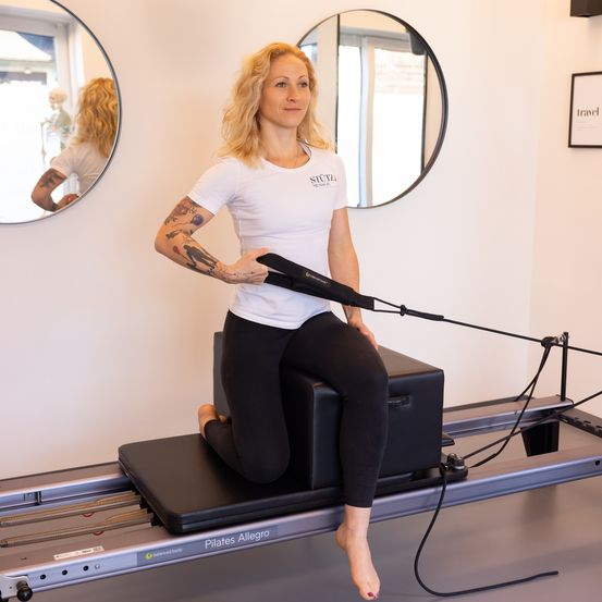 Eine Frau in einem weißen T-Shirt und schwarzen Leggings praktiziert Pilates auf einem Reformer und hält eine Schlaufe. Hinter ihr spiegelt ein Spiegel ihr Bild und ein Skelett. Ein Fenster ist im Hintergrund sichtbar.
