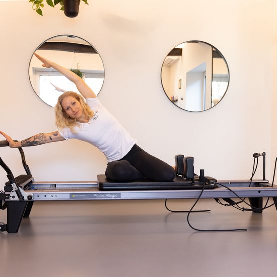 Eine Frau mit Tätowierungen praktiziert Yoga auf einer Pilates Allegro-Maschine in einem Raum mit zwei runden Spiegeln an der Wand.