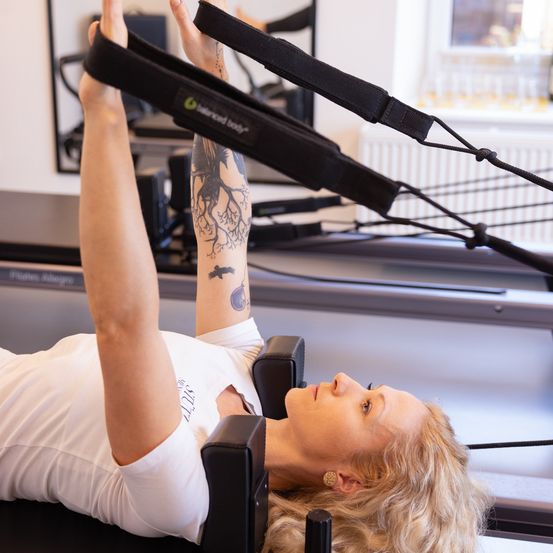 Eine Frau mit Tätowierungen an den Armen macht Pilates auf einem Reformer in einem Fitnessstudio.