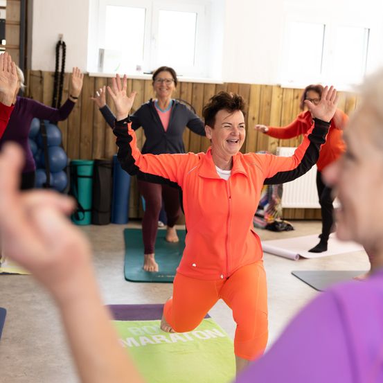 Eine Gruppe von Frauen praktiziert Yoga in einem Fitnessstudio. Eine Frau lächelt und hebt ihre Hände. Im Hintergrund befinden sich Yogamatten, ein Heizkörper und Hanteln.