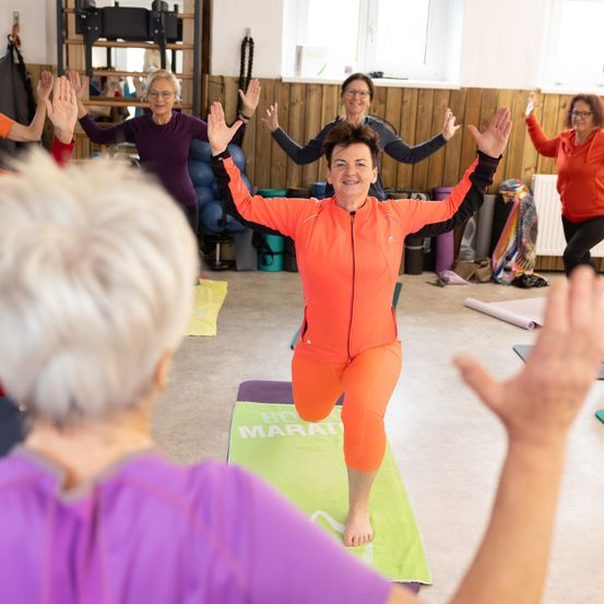 Eine Gruppe von Frauen nimmt in einem Studio an einer Yoga-Sitzung teil. Eine Frau in Orange führt, während andere folgen.