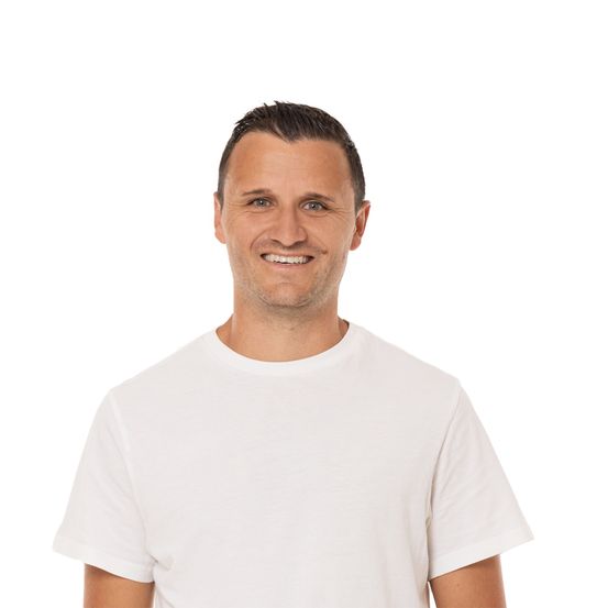 Ein Mann in einem weißen T-Shirt lächelt und schaut nach vorne. Er hat kurze Haare und steht vor einem schlichten weißen Hintergrund.