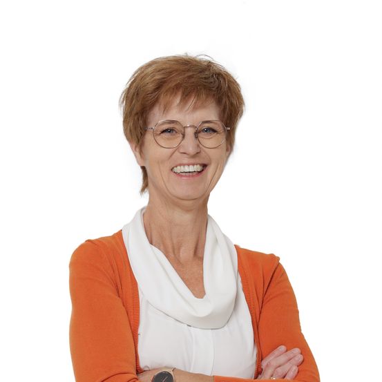 Eine lächelnde Frau mit kurzen Haaren, die eine Brille und einen orangefarbenen Cardigan trägt, posiert für ein Foto mit verschränkten Armen.