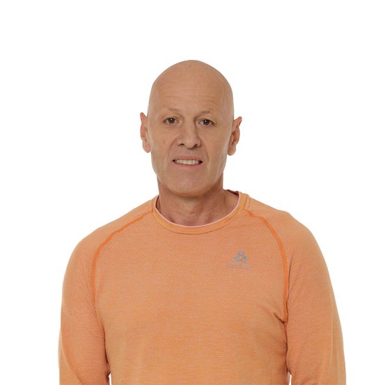 Ein kahlköpfiger Mann trägt ein orangefarbenes Langarmshirt mit einem Logo auf der linken Brust, lächelt und posiert für ein Foto.