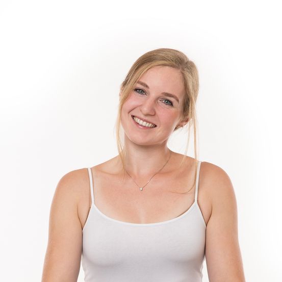 Eine Frau mit blonden Haaren lächelt und trägt ein weißes ärmelloses Oberteil. Sie steht vor einem weißen Hintergrund.