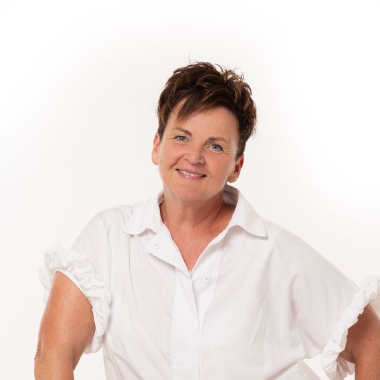 Eine Frau mit kurzen Haaren lächelt und posiert für ein Foto. Sie trägt ein weißes Hemd mit Rüschenärmeln.