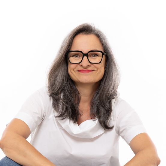 Eine Frau mit grauen Haaren, die eine Brille und ein weißes T-Shirt trägt, lächelt und sitzt auf einem weißen Hintergrund.