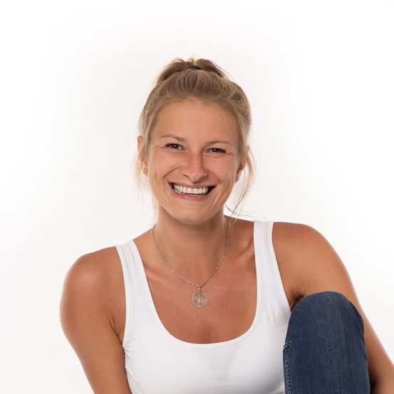 Eine Frau mit blonden Haaren im Pferdeschwanz, die ein weißes Tanktop und blaue Jeans trägt, lächelt fröhlich für ein Porträt.