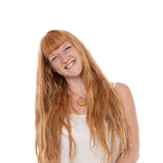 Eine Frau mit langen, welligen blonden Haaren und einem warmen Lächeln posiert für ein Porträt. Sie trägt ein weißes ärmelloses Oberteil und eine kreisförmige Anhängerkette.