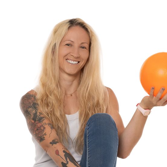 Eine Frau mit blonden Haaren und Tattoos am Arm lächelt und hält einen orangefarbenen Ball.