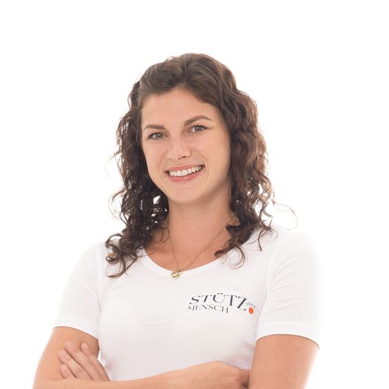 Eine lächelnde Frau mit lockigem Haar trägt ein weißes T-Shirt mit dem Text 'Stuetz Mensch', die Arme verschränkt.