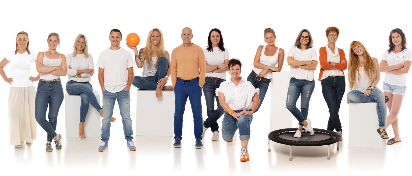 Eine Gruppe von sieben Erwachsenen, drei Männer und vier Frauen, posiert für ein Foto. Sie stehen und sitzen auf weißen Blöcken, eine Frau kniet und eine andere hält eine orange Kugel. Sie alle lächeln und scheinen in einem Studio zu sein.