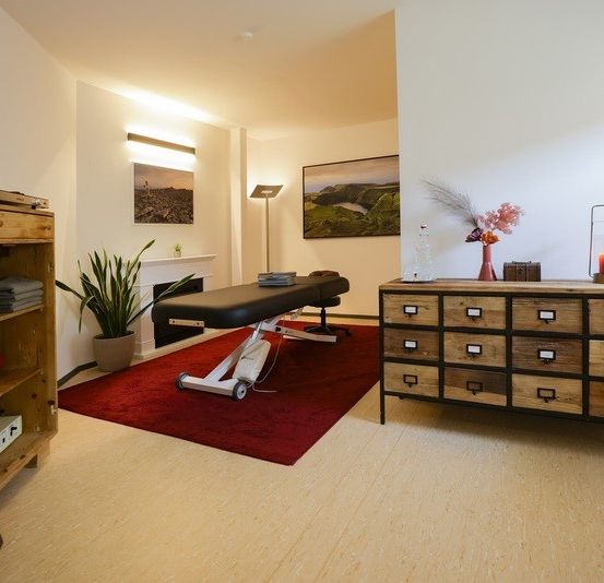 Ein gut beleuchteter Raum mit Holzböden verfügt über einen Massagetisch, einen roten Teppich und einen Schrank mit Schubladen. Ein Topf mit Pflanzen und eine Kerze verleihen dem Raum Atmosphäre.