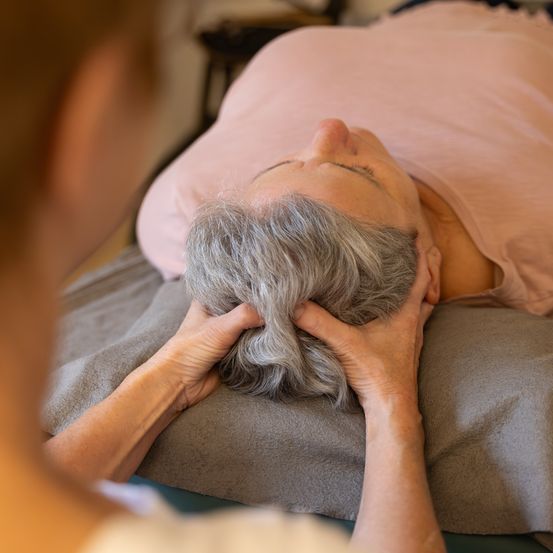 Eine ältere Frau liegt auf einem Bett, ihr Kopf ruht auf einem Kissen, während sie eine Massage von einer anderen Person erhält.