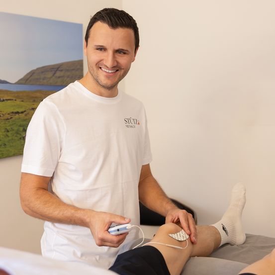 Ein Mann trägt einen Elektroden auf sein Knie auf, lächelt und schaut in die Kamera. Er trägt ein weißes T-Shirt und schwarze Shorts.