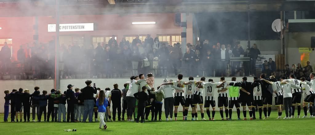 A soccer team in black and white uniforms is huddled together on a field with smoke and spectators behind them.