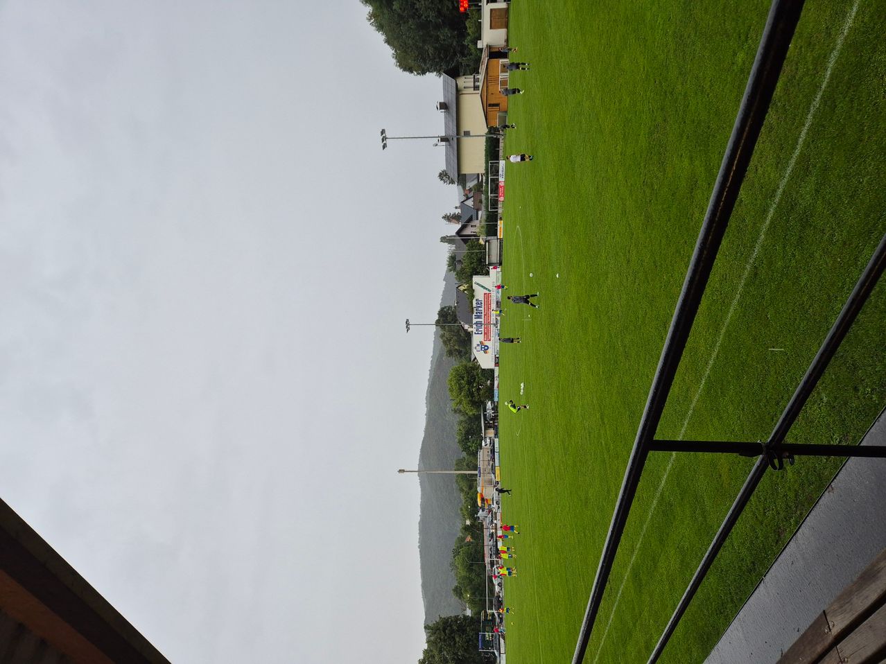 Ein Fußballfeld mit grünem Rasen und verstreuten Spielern unter einem bewölkten Himmel. Es gibt Straßenlaternen und ein Gebäude in der Ferne.