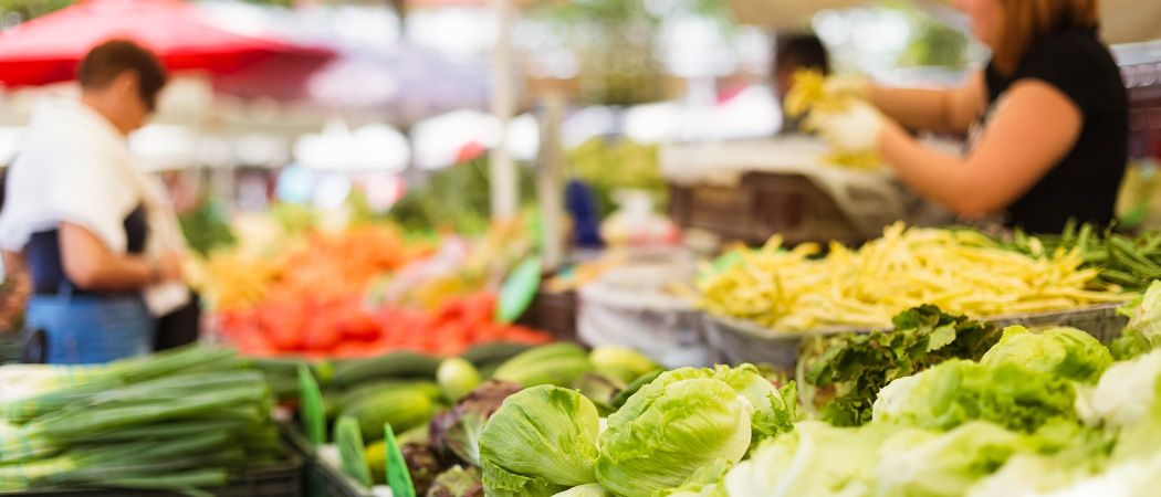 Ein Markt im Freien mit verschiedenen Gemüsesorten wie Tomaten, Salat und Gurken, die unter roten Schirmen ausgestellt sind.