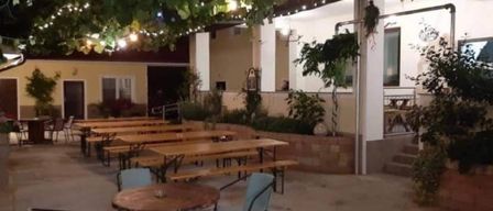 A patio with wooden tables and benches, surrounded by green foliage. Decorative lights hang from above. The setting appears to be an outdoor restaurant or cafe.