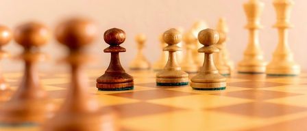 Ein Schachbrett mit Holzfiguren. Der König steht in der Mitte, flankiert von Bauern. Der Hintergrund ist rosa und verschwommen.