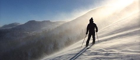 Ein Skifahrer fährt den schneebedeckten Hang hinunter, wobei die Sonne die Szene beleuchtet und Schatten auf den Schnee wirft.