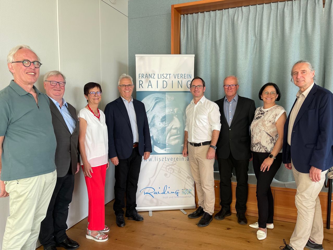 Sieben Personen stehen vor einem Banner mit dem Text Franz Liszt Verein Raiding. Sie alle lächeln und scheinen für ein Foto zu posieren.